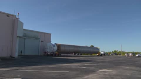 A wide shot shows a truck pulling a long rocket booster near a hangar.