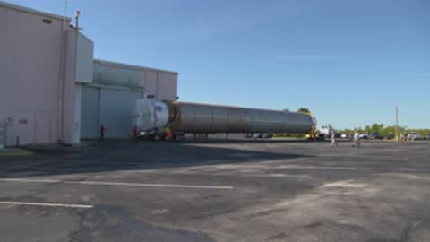 A wide shot shows a truck pulling a rocket booster toward a large hangar.