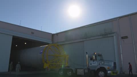 A wide shot shows a truck pulling a rocket booster backward into a hangar.