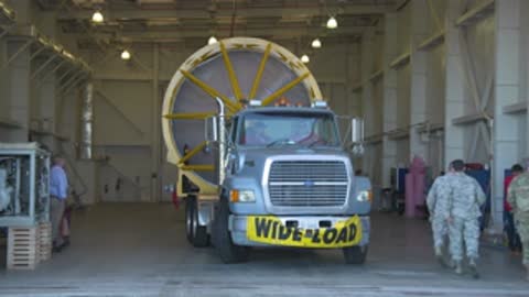 A medium shot shows a truck with a wide load sign parked inside a hangar.