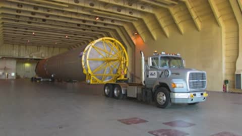 Medium tracking shot of a semi-truck transporting a large rocket stage indoors.