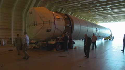 Medium shot of workers walking around a large rocket component inside a hangar.