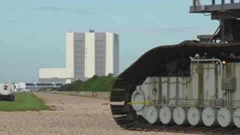 Close-up of crawler treads moving along gravel road with VAB in background.