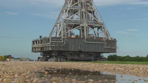 Medium shot of crawler-transporter moving toward camera on gravel roadway.