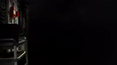 Wide shot inside a dark equipment room with server racks and a person working.