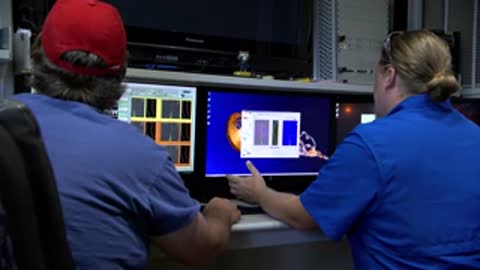 Medium shot of two people sitting at computer terminals monitoring weather simulation data on multiple screens.