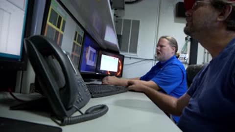 Medium shot of two men seated at computer stations, one looking up toward another screen.
