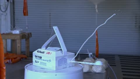Close-up of a radiosonde weather sensor box with a long wire antenna on a table.
