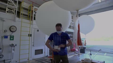 Medium shot of a person wearing a mask handling equipment near a large weather balloon indoors.