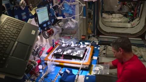 An astronaut in a red shirt works with equipment modules on a table in the station.