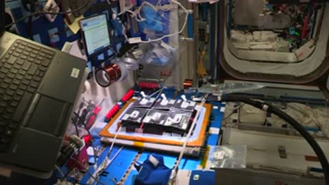 A hand in a blue glove reaches across a table inside the space station module.