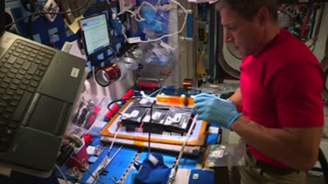 An astronaut in a red shirt holds a tool over a workspace inside the space station.