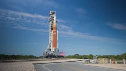 A white car drives on the road in front of the SLS rocket.