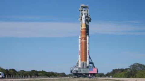 The SLS rocket moves slowly down the crawlerway under a blue sky.
