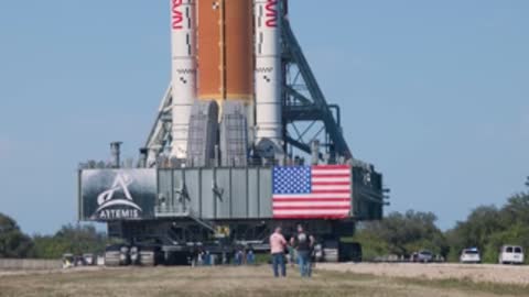 People stand near the crawler platform holding the SLS rocket outdoors.