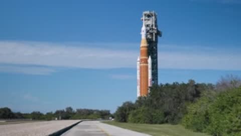 The SLS rocket passes behind green trees along the crawlerway path.