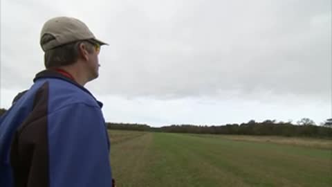 A man in a blue jacket and cap watches the aircraft in flight.