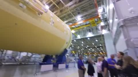 Camera pans along a large yellow rocket component inside a facility.
