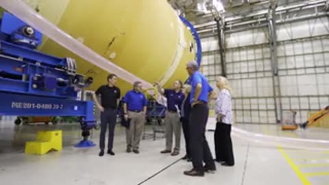 People standing around a large yellow rocket section inside a warehouse.