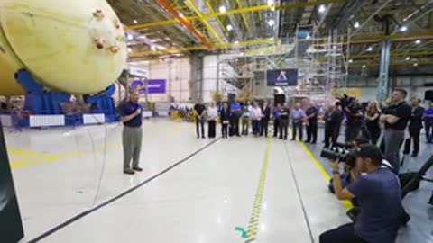 A man speaking to a group inside a large warehouse near a rocket part.