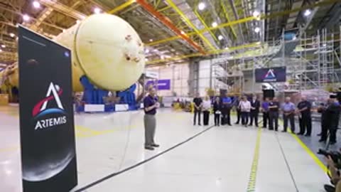 A wide indoor shot of a speaker addressing an audience near a rocket part.
