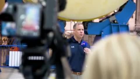 A man speaking into a microphone seen through camera equipment and a crowd.