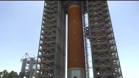 Camera pans down a tall, orange rocket structure outdoors on a sunny day.