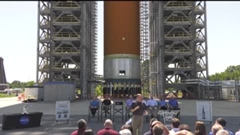 People seated outdoors watching a presentation in front of a rocket model.