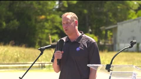 A man speaking into a microphone outdoors, gesturing with his hands.