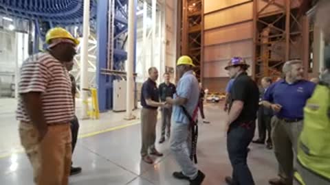 People walking around inside a large industrial facility with yellow safety helmets.