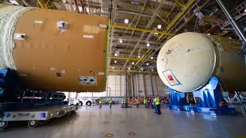 Medium shot inside a workshop as two large cylindrical rocket sections approach each other.
