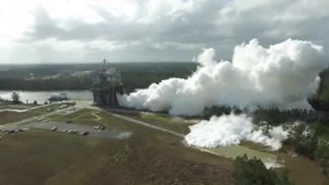 Aerial tracking shot of white smoke billowing from a rocket test stand near trees.