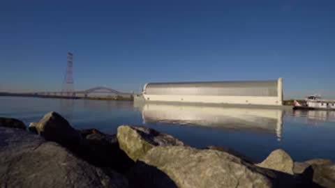 Wide tracking shot of a covered barge carrying a large structure along a river.