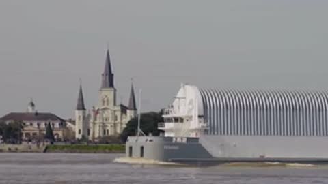 Medium tracking shot of a covered barge passing a cathedral and buildings along a riverbank.
