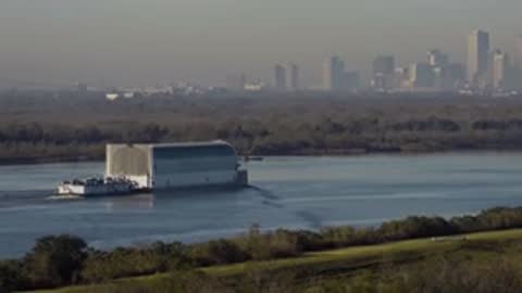Wide tracking shot of a covered transport barge on a river with a distant city skyline.
