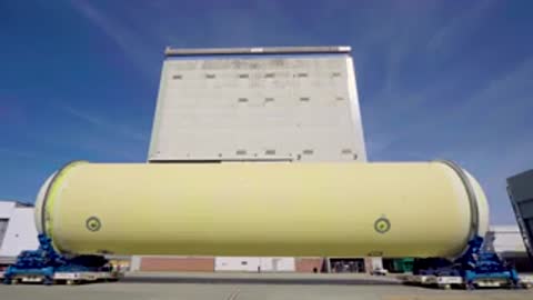 Medium tracking shot of a large cylindrical rocket part outside near a building.