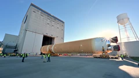 Transport vehicle carrying rocket stage past large industrial building