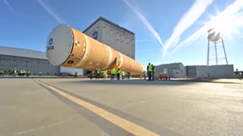 Rocket stage transport moving past water tower and building