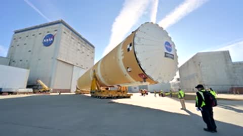 Rocket stage moving on transport vehicle past industrial buildings