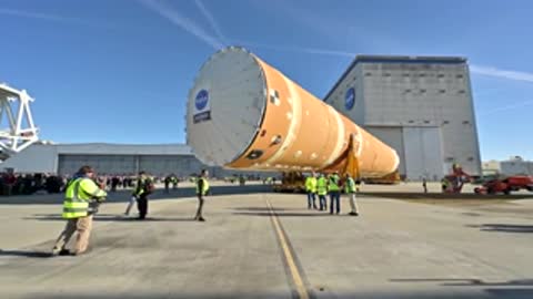 Workers and rocket stage moving across paved area outdoors