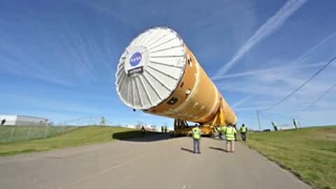 Rocket stage transport moving along paved path near grassy area