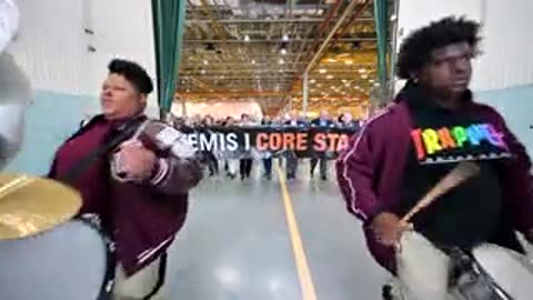 Marching band members walking and playing instruments indoors