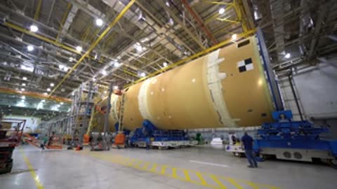 Wide view of a long horizontal rocket component inside a large factory.