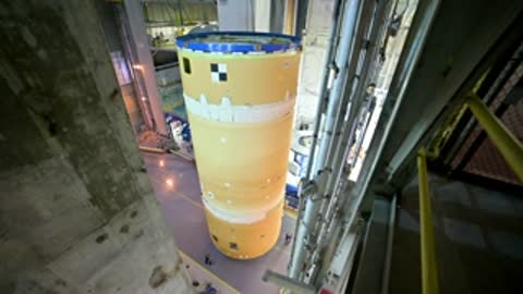 Vertical cylindrical rocket component standing upright inside a large facility.