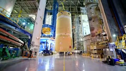 Long cylindrical rocket component standing vertically inside a large manufacturing facility.