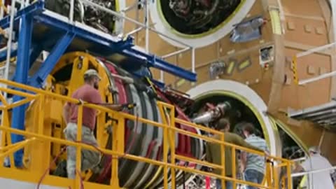 Workers assembling rocket engine parts on a large structure indoors.