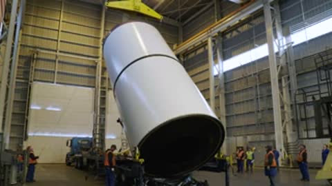 Large white cylindrical rocket component being lifted by crane indoors.