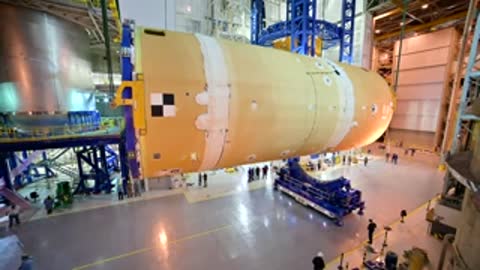 Horizontal cylindrical rocket component being lifted by a crane in a facility.