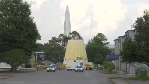 Launch vehicle stage adapter transported along road past tree-lined area.