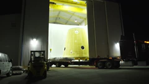 A truck hauling a yellow component backs into a large building at night.